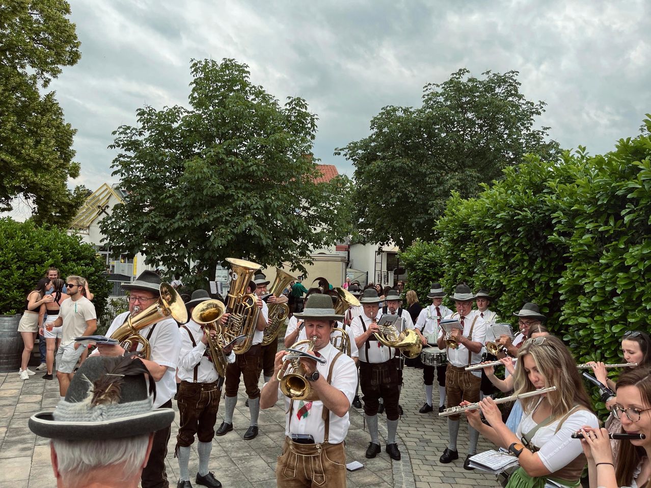 Eine Blaskapelle spielt im Freien, mit Musikern in traditioneller bayerischer Kleidung, einschließlich Hüten und Hosenträgern, die verschiedene Instrumente wie Trompeten, Posaunen und Tuben spielen. Der Ort ist eine von Bäumen und Gebäuden gesäumte Straße, mit einem bewölkten Himmel darüber.