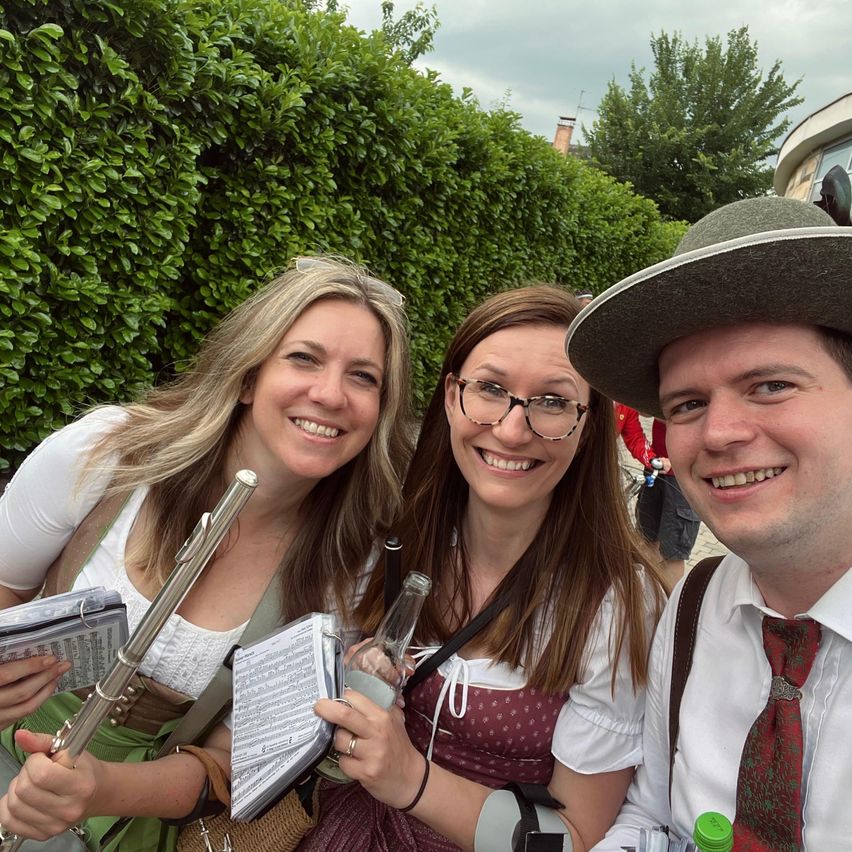 Drei Menschen lächeln in einem Park und halten Musikinstrumente. Eine Frau hat eine Flöte, eine andere eine Flasche und ein Buch, und der Mann trägt einen Hut und eine Krawatte. Hinter ihnen steht eine Hecke und ein Gebäude.