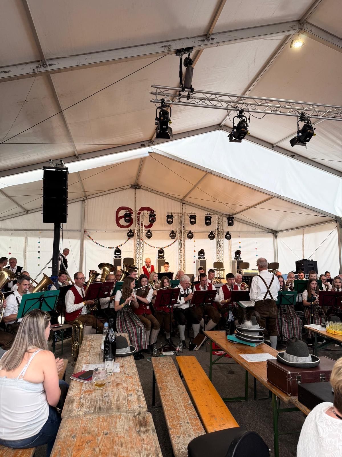 Eine Band spielt in einem Zelt im Innenraum mit verschiedenen Instrumenten und sitzenden Zuhörern. Bühnenbeleuchtung ist vorhanden.