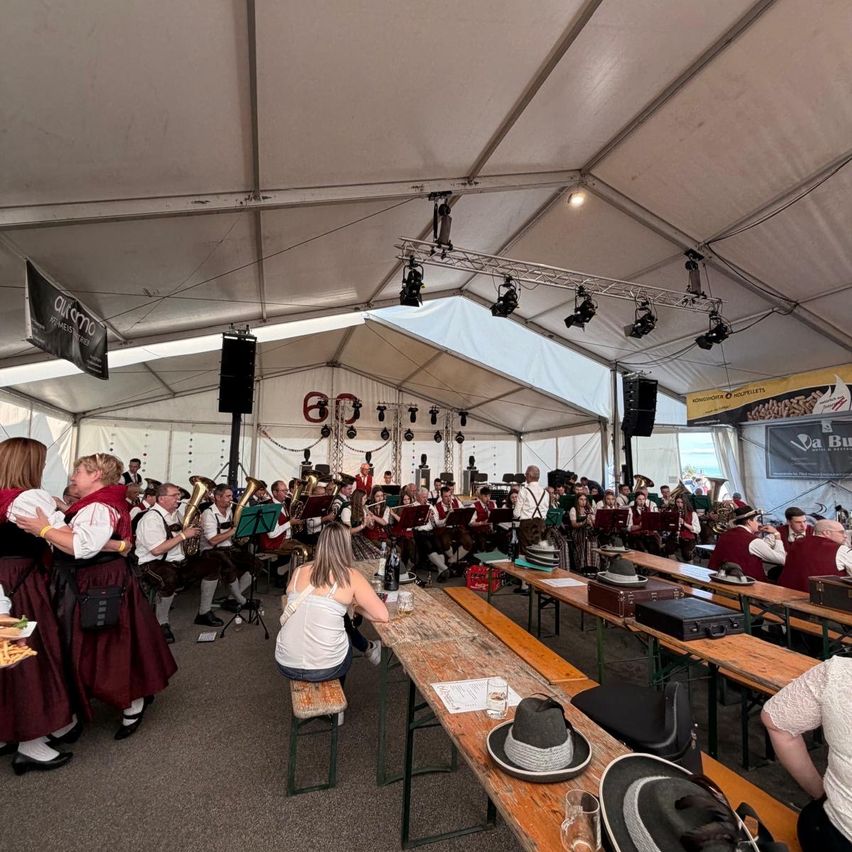 Ein Blasorchester spielt in einem Zeltpavillon, während eine Menge Menschen um Tische herumsitzt, zuhört und die Musik genießt.