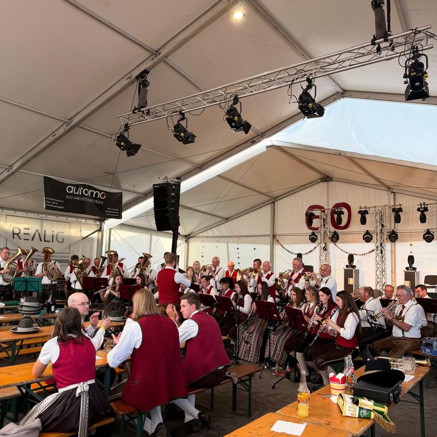 Ein Orchester spielt unter einem großen weißen Zelt. Musiker sitzen an Tischen, während andere im Hintergrund stehen. Das Publikum sitzt vorne und genießt die Musik.