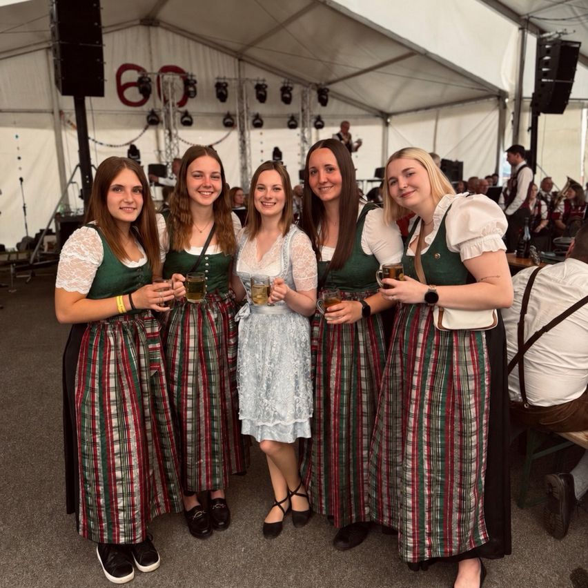 Fünf Frauen, gekleidet in traditioneller deutscher Kleidung, halten Getränke in einem festlichen Zelt. Eine Band spielt im Hintergrund, und mehrere Leute sitzen auf Bänken.