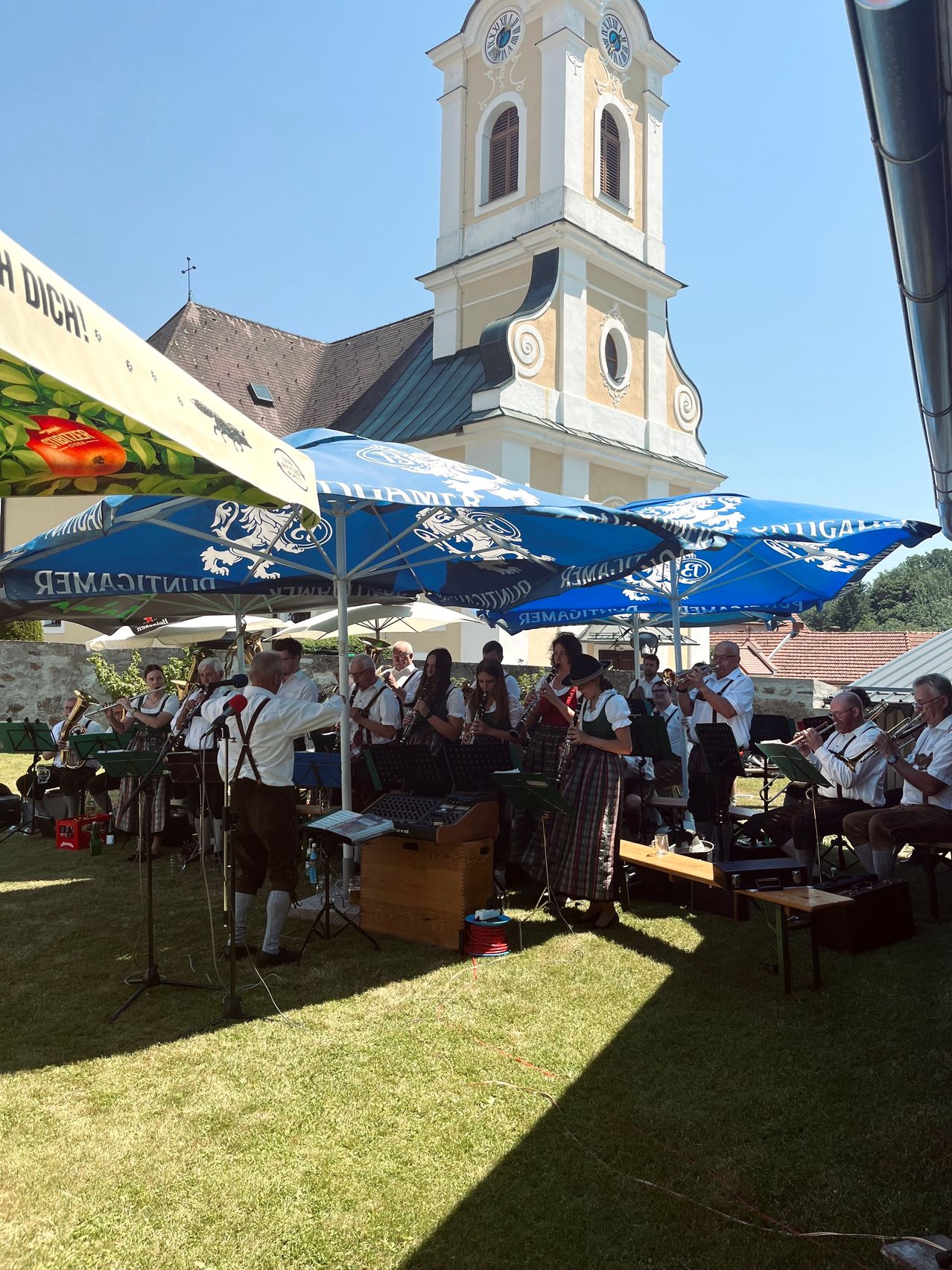 Eine Gruppe Musiker tritt unter großen Schirmen vor einem historischen Gebäude im Freien auf. Sie spielen verschiedene Instrumente, und einige sitzen auf Bänken.