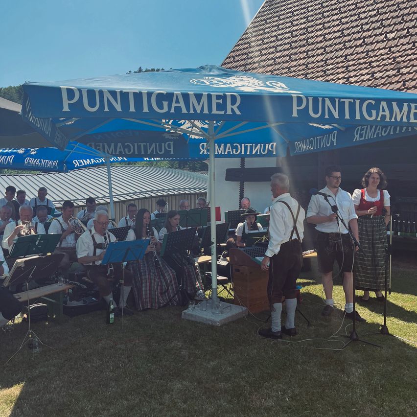Ein Musikensemble spielt Instrumente und singt unter einem blauen Zelt, während Leute sitzen und stehen, in einer sonnigen Freiluftumgebung.