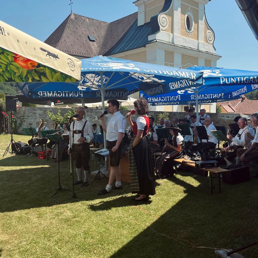 Eine Musikband spielt auf einem Rasenfeld unter blauen Sonnenschirmen. Zwei Personen singen und halten Mikrofone in der Hand, während andere Musikinstrumente spielen. Hinter ihnen ist ein großes Gebäude mit einem Turm zu sehen.