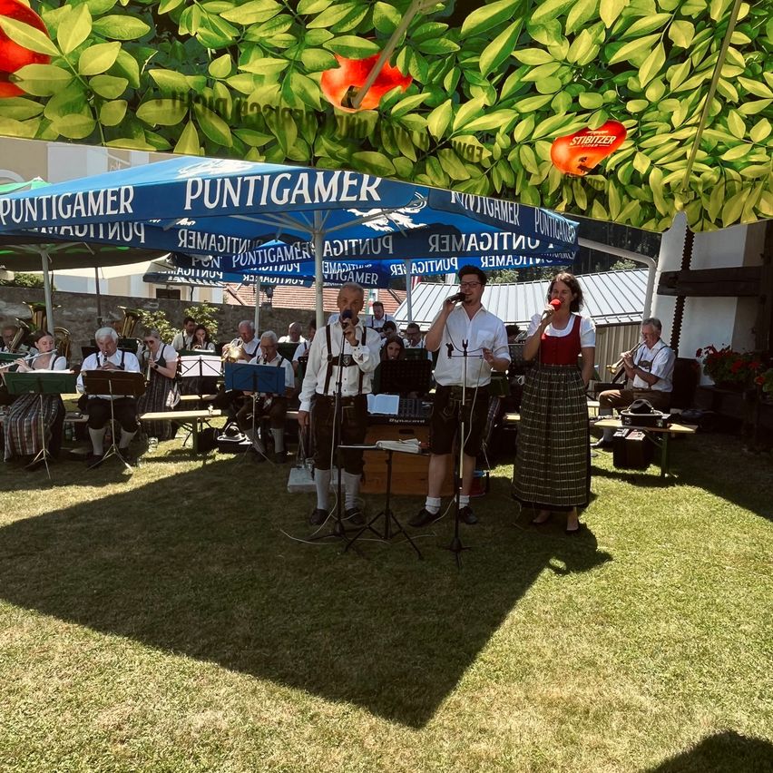 Eine Gruppe von Musikern tritt auf einem Rasenfeld unter einem blauen Zelt mit Puntigamer auf. Eine Frau und zwei Männer singen und halten ein Mikrofon.