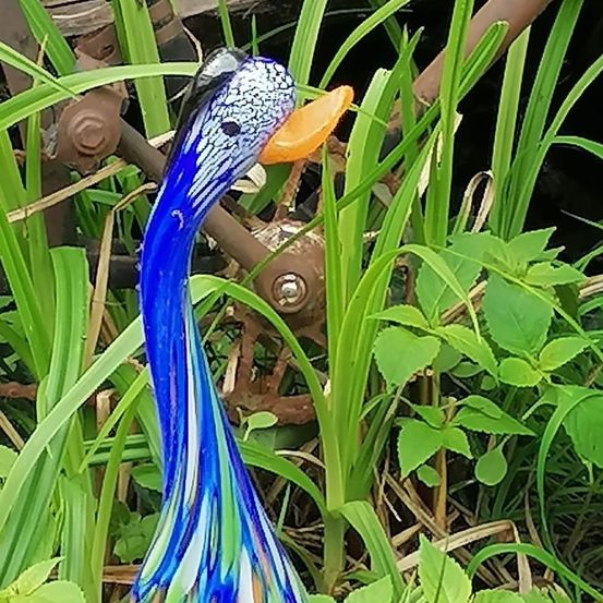 Eine blaue Glasentenskulptur mit einem leuchtend orangefarbenen Schnabel ist teilweise von hohem, grünem Gras verdeckt. Ein Metallrad ist teilweise hinter der Ente sichtbar.