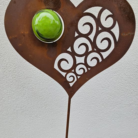 Eine herzförmige Metallskulptur mit einem grünen Glaszentrum und komplizierten Wirbelmustern. Montiert an einer weißen Wand.