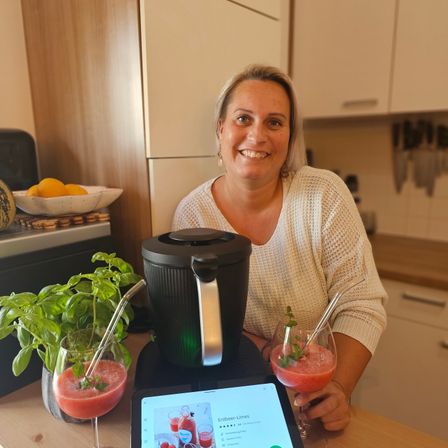 Eine Frau lächelt in einer Küche neben einem Mixer mit zwei Getränken. Eine Pflanze und eine Schüssel mit Zitronen sind im Hintergrund.