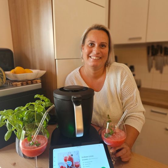Eine Frau lächelt in einer Küche neben einem Mixer mit zwei Getränken. Eine Pflanze und eine Schüssel mit Zitronen sind im Hintergrund.
