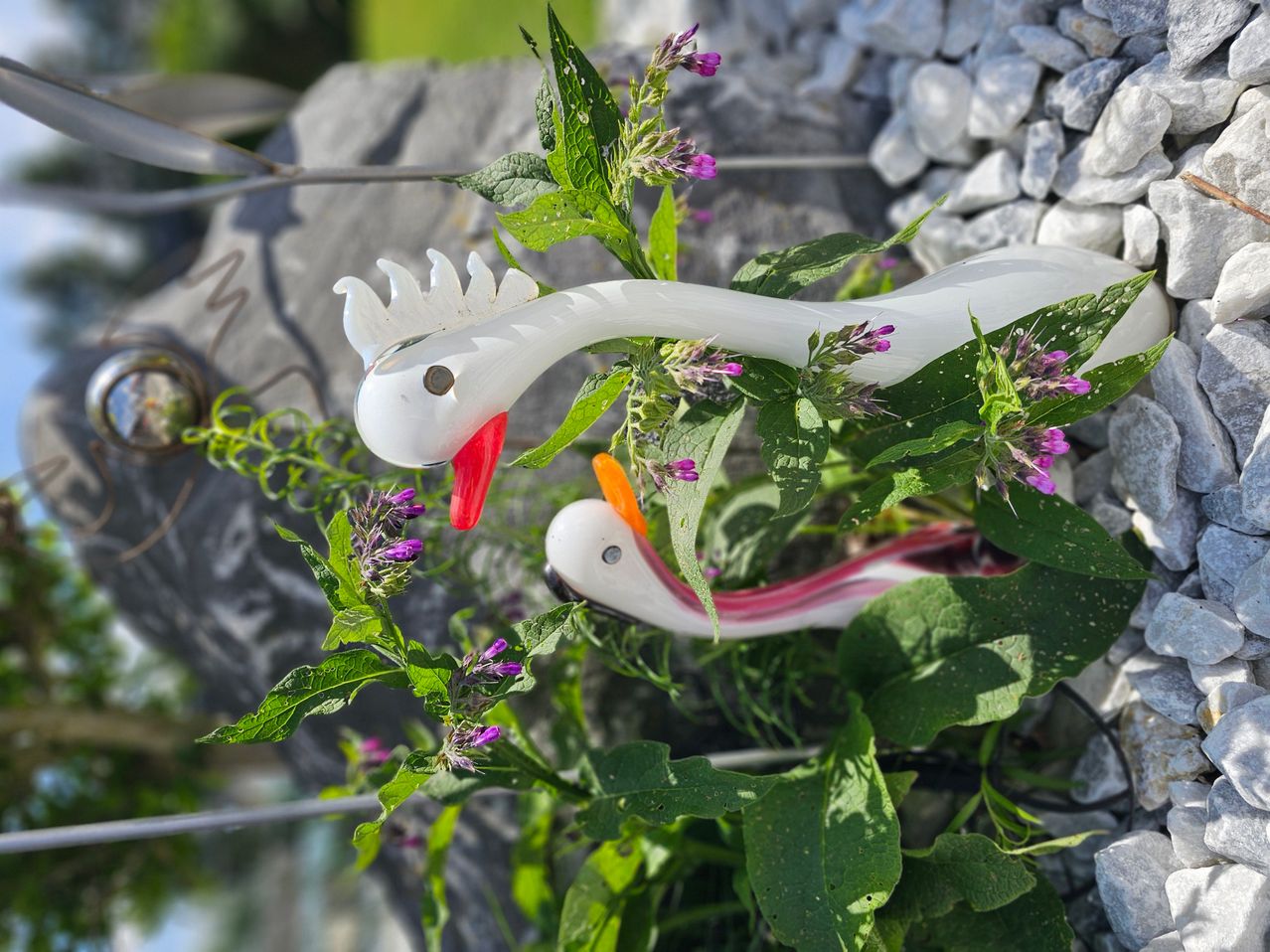 Zwei Glasschwäne mit roten Schnäbeln sitzen auf üppigen grünen Pflanzen mit lila Blüten, vor dem Hintergrund grauer Steine und Kiesel.