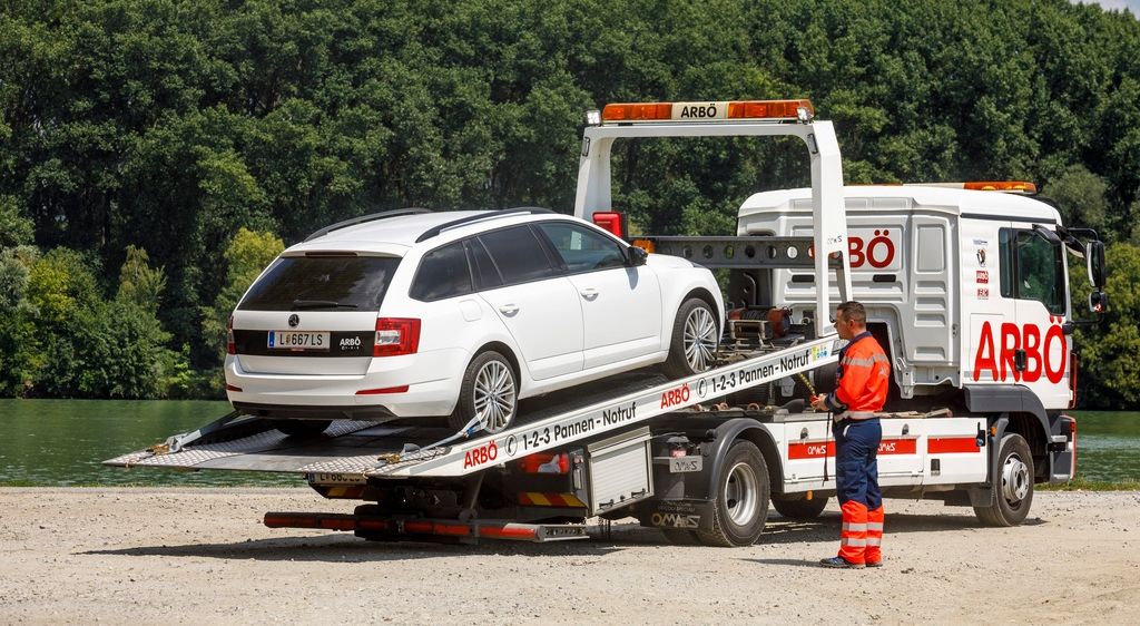 Ein weißes Auto wird von einem Abschleppwagen abtransportiert. Der Lastwagen hat ein Nummernschild, das ARBO liest. Das Auto hat ein Nummernschild, das L 667 LS liest.