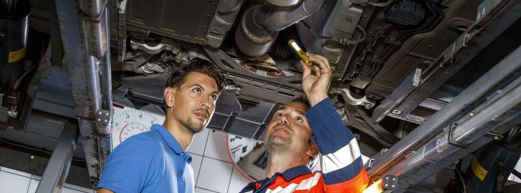Zwei Männer in einem Autoservice, einer hält eine Taschenlampe und untersucht die Unterseite eines Autos. Im Hintergrund ist eine Uhr sichtbar.