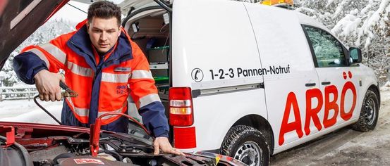 Ein Mann in einer orangefarbenen Uniform repariert ein Auto. Er benutzt ein Werkzeug an der Batterie. Der Autohauben ist offen. Hinter ihm steht ein weißer Lieferwagen mit einem roten Schild, das ARBO 1-2-3 besagt.