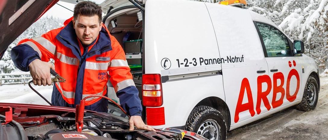 Ein Mann in einer orangefarbenen Uniform repariert ein Auto. Er benutzt ein Werkzeug an der Batterie. Der Autohauben ist offen. Hinter ihm steht ein weißer Lieferwagen mit einem roten Schild, das ARBO 1-2-3 besagt.