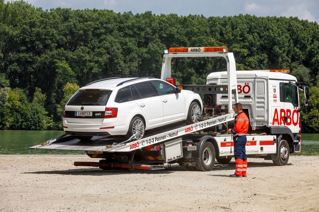 Ein weißer Wagen wird auf einem Flachbett-Lkw transportiert. Der Fahrer steht neben dem Lastwagen und trägt eine orange Weste. Bäume sind im Hintergrund.