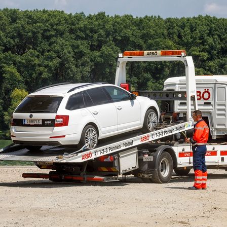 Ein weißer Wagen wird auf einem Flachbett-Lkw transportiert. Der Fahrer steht neben dem Lastwagen und trägt eine orange Weste. Bäume sind im Hintergrund.