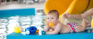 Ein Baby spielt in einem kleinen Pool mit Spielzeug, während jemand einen Wassersprüher in der Nähe hält. Der Pool ist blau und hat eine Fontäne.