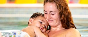 Eine Mutter hält ihr Baby im Schwimmbecken, beide lächeln vor Freude, umgeben von Wasserspritzern.