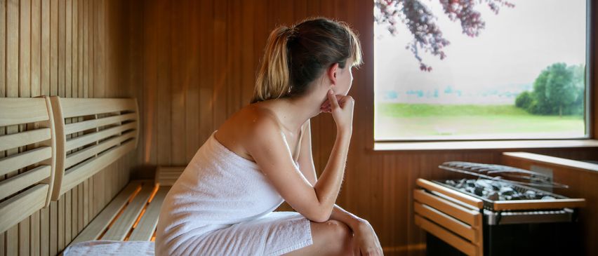 Eine Frau in einem weißen Handtuch sitzt in einer Holzsauna und schaut durch das Fenster auf einen Baum.