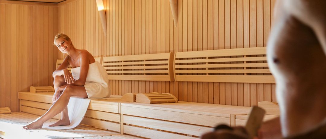 Ein Mann und eine Frau in einer Holzsauna. Die Frau sitzt auf einer Bank und lächelt, während der Mann vor ihr steht und einen Holzeimer hält.