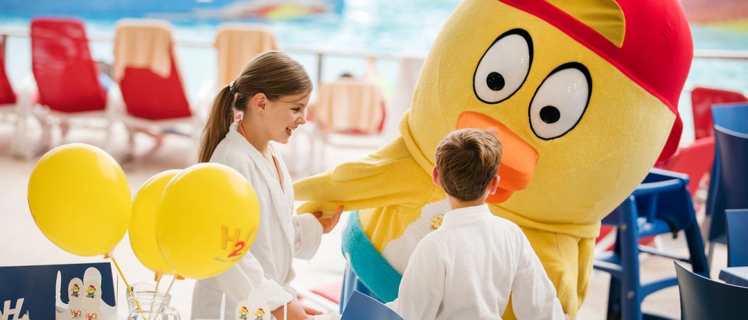 Zwei Kinder in weißen Gewändern interagieren mit einem Maskottchen bei einer Indoor-Pool-Party mit gelben Luftballons und einem festlichen Tisch.