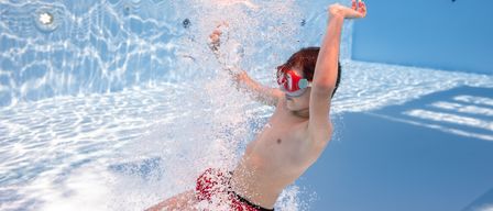 Ein junger Junge schwimmt unter Wasser in einem Pool mit Schwimmbrille und roten Badeshorts, wobei Wasser um ihn herumspritzt.