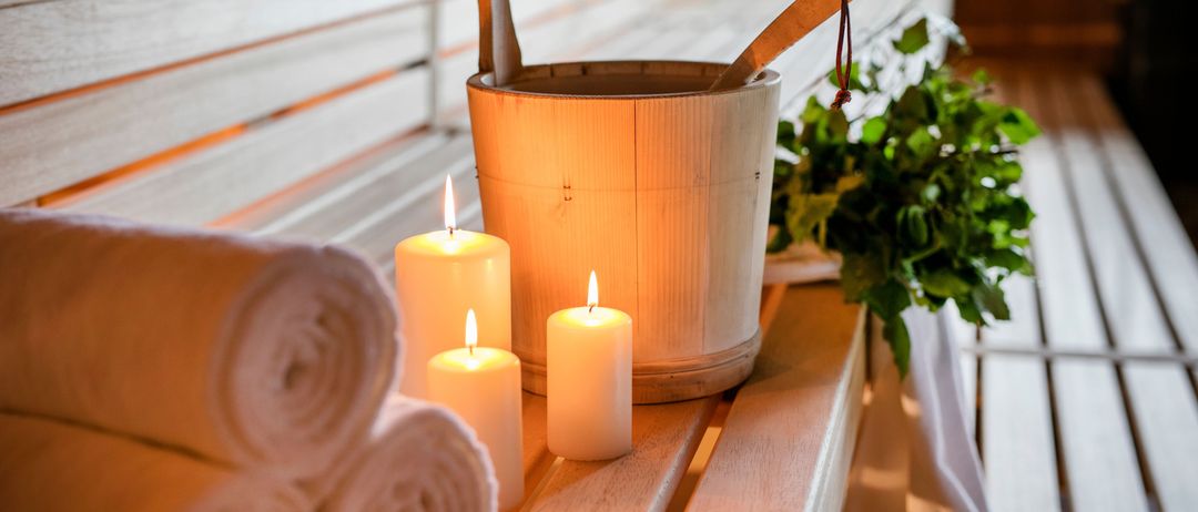 Eine Holzbank in einer Sauna mit einem Eimer, einem Schöpflöffel, zwei brennenden Kerzen und gerollten Handtüchern.