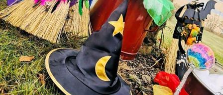 Eine Halloween-Dekoration mit einem Hexenhut, Besen und einem Kürbis-Eimer auf einem Grasfeld mit einem Baum und einem Gebäude im Hintergrund.