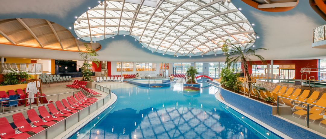 Ein großer Innenpool mit einer Glaskuppeldecke, umgeben von roten Liegestühlen und Grünpflanzen. Der Pool ist sauber und gut gepflegt.
