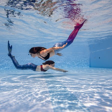 Zwei Frauen schwimmen mit Meerjungfrauschwänzen unter Wasser in einem Pool. Die Frau vorne hat einen violetten Schwanz und die Frau dahinter einen blauen Schwanz. Der Pool ist blau mit einer blauen Wand an der Seite.