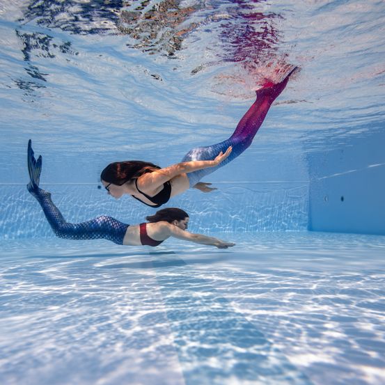 Zwei Frauen schwimmen mit Meerjungfrauschwänzen unter Wasser in einem Pool. Die Frau vorne hat einen violetten Schwanz und die Frau dahinter einen blauen Schwanz. Der Pool ist blau mit einer blauen Wand an der Seite.