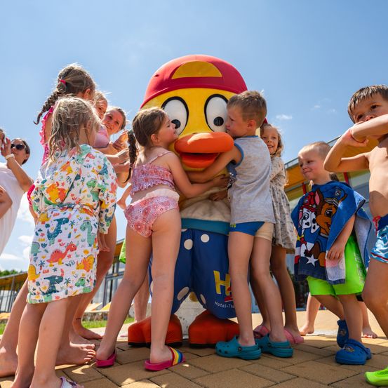 Eine Gruppe von Kindern in Badeanzügen umarmt eine große, bunte Zeichentrickfigur an einem sonnigen Tag.