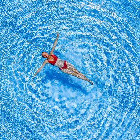 Eine Frau schwimmt im Pool und streckt ihre Arme aus, wodurch eine Welle in dem blauen Wasser entsteht.