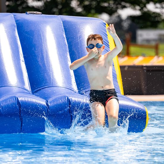 Ein Junge mit Schwimmbrille springt in eine blaue aufblasbare Wasserrutsche in einem Freibad.