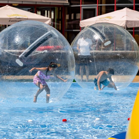 Zwei Kinder spielen in großen transparenten Bällen im Pool, eines springt und das andere kriecht darin.