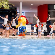 In einem Innenpool unterhält ein Maskottchen in einem gelben Kostüm Kinder, während Erwachsene zusehen. Der Pool ist mit Wasser gefüllt und von weißen Stühlen umgeben.