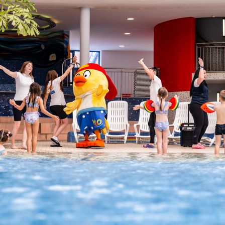 In einem Innenpool unterhält ein Maskottchen in einem gelben Kostüm Kinder, während Erwachsene zusehen. Der Pool ist mit Wasser gefüllt und von weißen Stühlen umgeben.