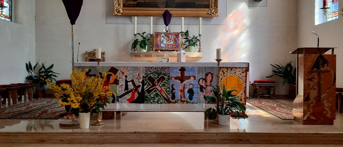 Ein Kirchenaltar ist mit einem farbenfrohen Kunstwerk, einem Kreuz und Kerzen geschmückt. Ein Bild hängt an der Wand und eine Pflanze steht auf dem Altar.