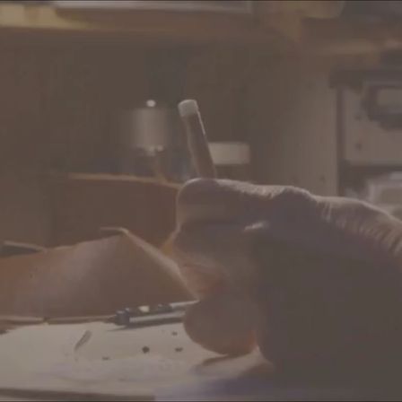 Eine Hand hält einen Stift über ein Stück Papier auf einem Schreibtisch. Ein brauner Lederbeutel und ein verschwommener Topf im Hintergrund.