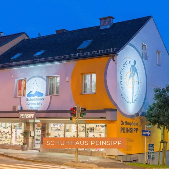 Die Vorderseite von Schuhhaus Peinsiipp, einem orthopädischen Schuhgeschäft, ist nachts beleuchtet. Das Gebäude hat große Fenster, die verschiedene Schuhe präsentieren. Das Schild trägt den Namen Schuhhaus Peinsiipp und in der Nähe befindet sich eine Ampel.