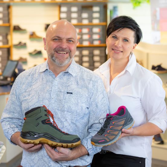 Ein Mann und eine Frau stehen in einem Schuhgeschäft und lächeln. Sie halten jeweils ein Paar Wanderschuhe, eines grün und eines pink.