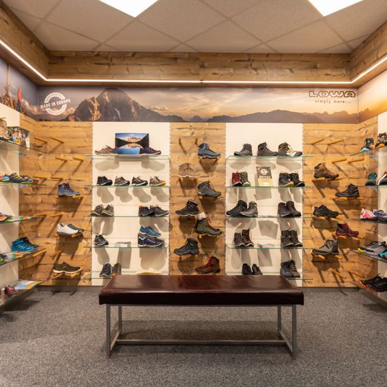 Ein Schuhgeschäft zeigt verschiedene Wanderschuhe auf Glasregalen. Die Wände sind aus Holz und eine Bank steht in der Mitte. Ein Berg und ein Sonnenuntergang sind auf einem Banner darüber abgebildet.