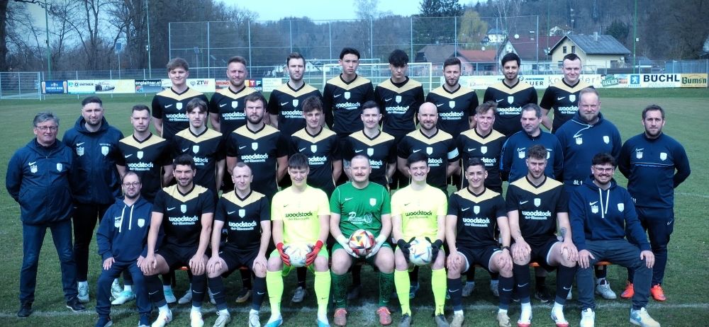 Eine Fußballmannschaft in gelben und schwarzen Trikots posiert für ein Foto auf einem Feld. Die Spieler halten Fußballbälle. Im Hintergrund befinden sich ein Zaun, Bäume und Häuser.