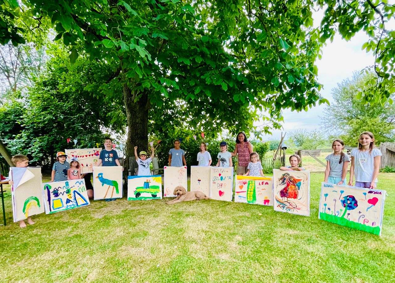Eine Gruppe von Kindern und Erwachsenen steht in einem Grasbereich unter einem Baum, jeder hält ein farbenfrohes Gemälde. Ein Hund liegt in der Nähe auf dem Boden.