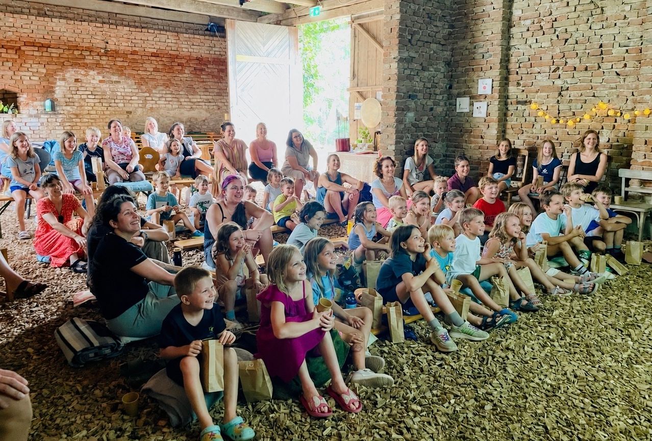 Eine Gruppe von Erwachsenen und Kindern sitzt auf dem Boden in einem Raum mit Ziegelwänden und schaut etwas an. Sie halten Papiertüten und einige tragen Sandalen.