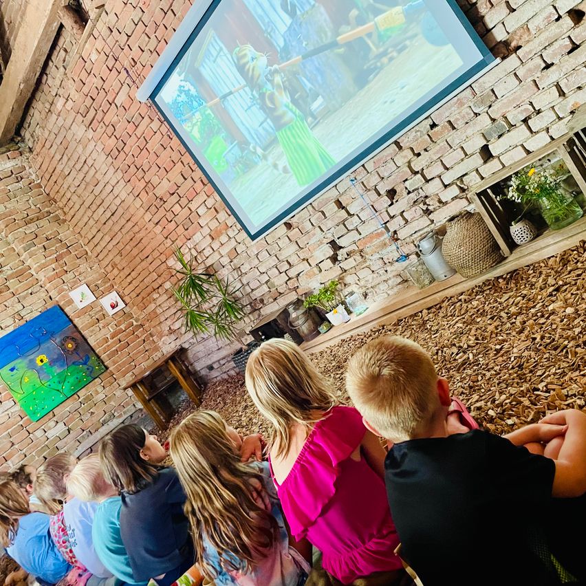 Kinder lauschen einem Film auf einer Leinwand in einem rustikalen Raum mit Ziegelwänden.