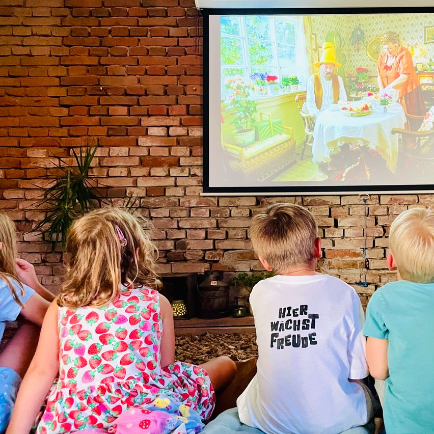 Vier Kinder sitzen vor einer Projektionsleinwand, die einen Cartoon zeigt. Der Junge in der Mitte trägt ein weißes T-Shirt mit deutschem Text.