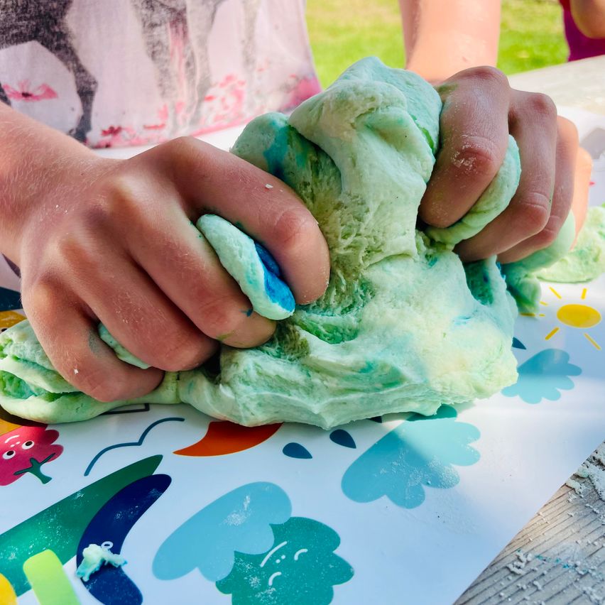 Ein Kind spielt mit grünem und blauem Schleim auf einem Tisch mit einem farbenfrohen Blumendesign.