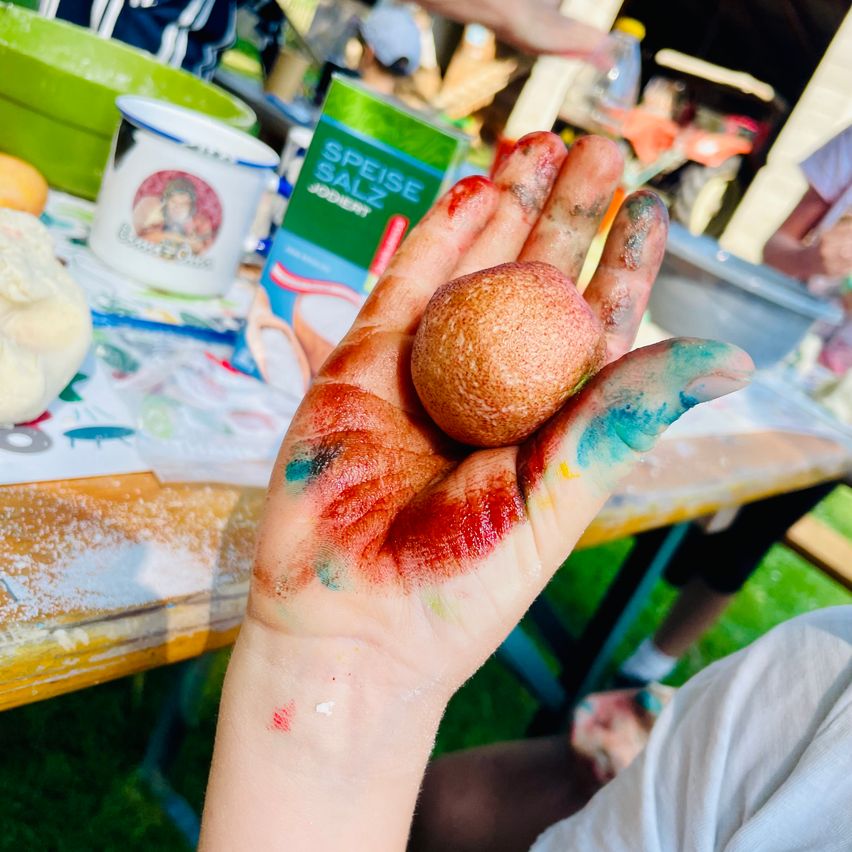 Eine mit bunter Farbe bedeckte Hand hält ein kleines, rundes Objekt, möglicherweise einen Ball, mit roter, blauer und gelber Farbe darauf. Der Tisch dahinter hat verschiedene Gegenstände, darunter eine Tasse, eine Schüssel und eine Box mit Gewürzen.
